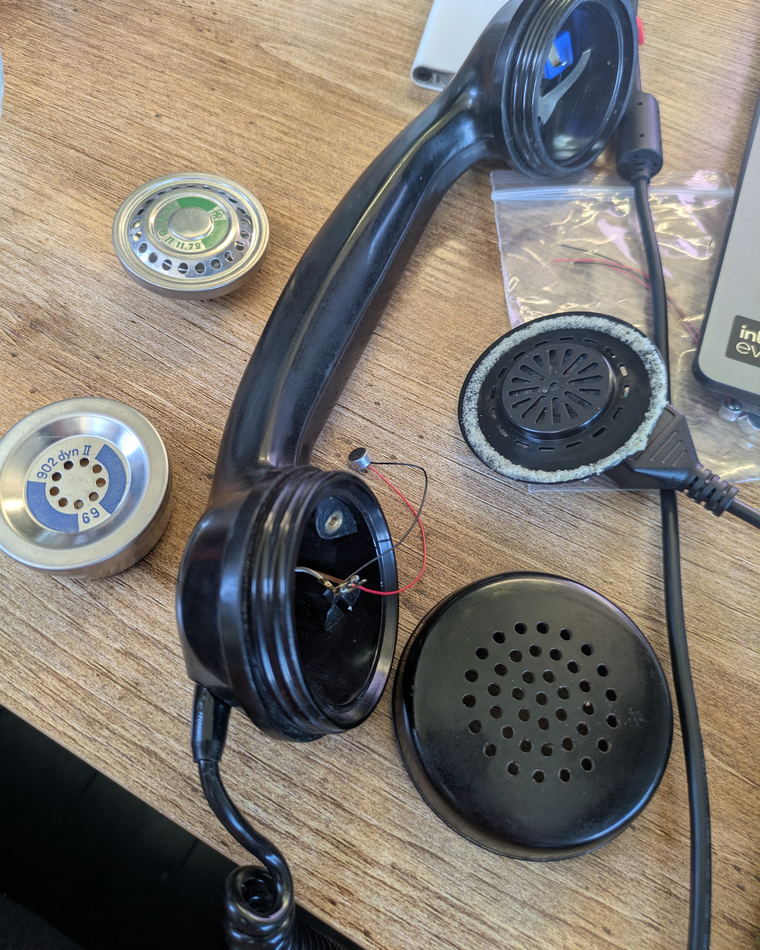 An opened telephone handset on a workbench beside its two original round metal capsules and a black speaker grille.