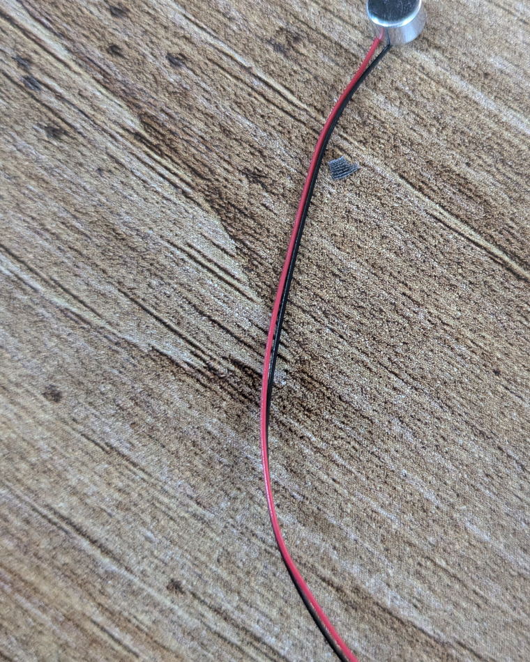 A very small cylindrical microphone capsule on a wooden surface, with a thin red and black wire pair running from it.