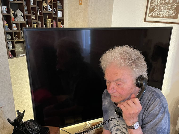 An elderly man with white curly hair sitting in his living room, holding the handset of a black rotary telephone to his ear.