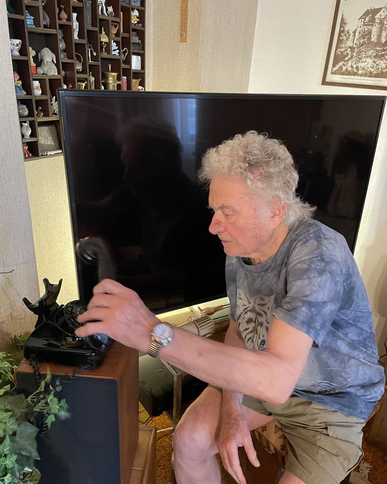 The elderly man reaching out and putting his hand on the handset of the telephone on the cabinet beside him.