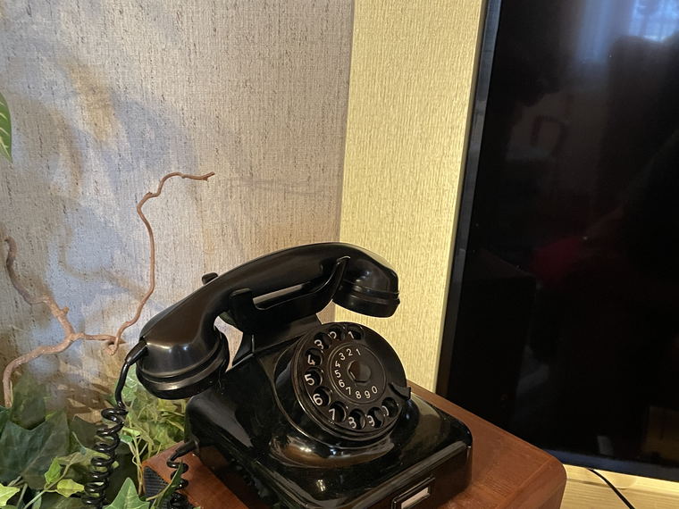 The black rotary telephone standing on a wooden cabinet in a living room, in front of a large switched-off television.