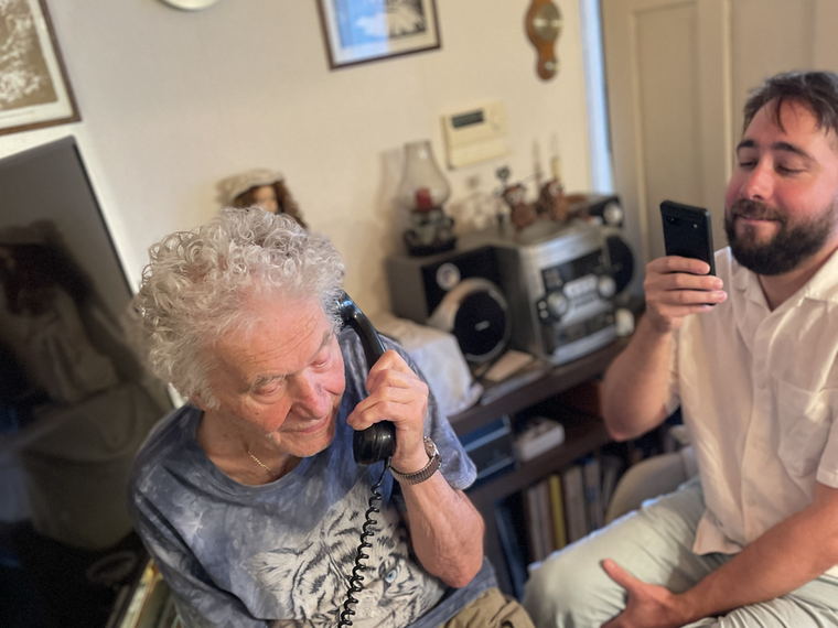 The elderly man on the telephone while the younger man beside him holds up a mobile phone.