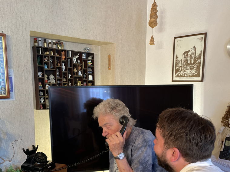The elderly man on the telephone with a younger bearded man sitting close beside him, leaning in to listen.