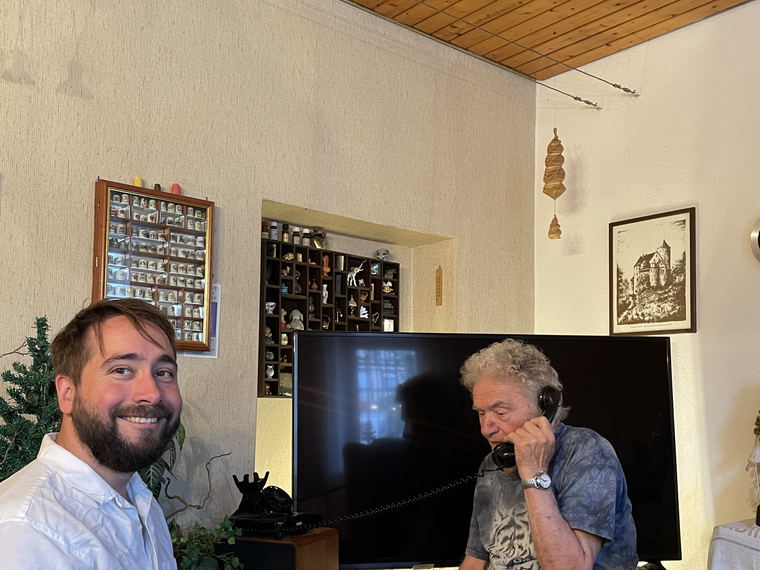 A wider view of the living room: the younger man smiling on the left, the elderly man on the telephone on the right.