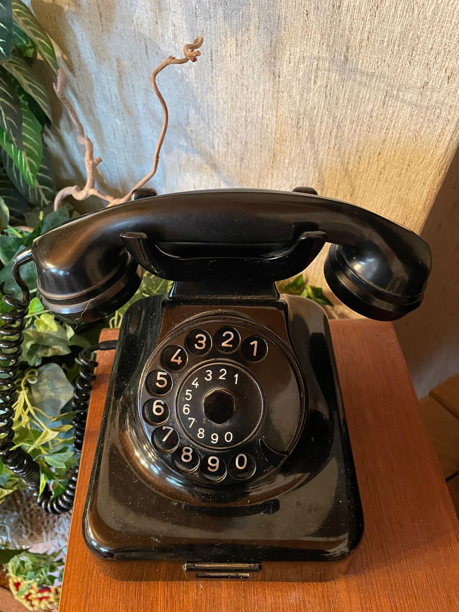 A black bakelite rotary telephone with its handset resting on the cradle, standing on a teak sideboard beside a houseplant.