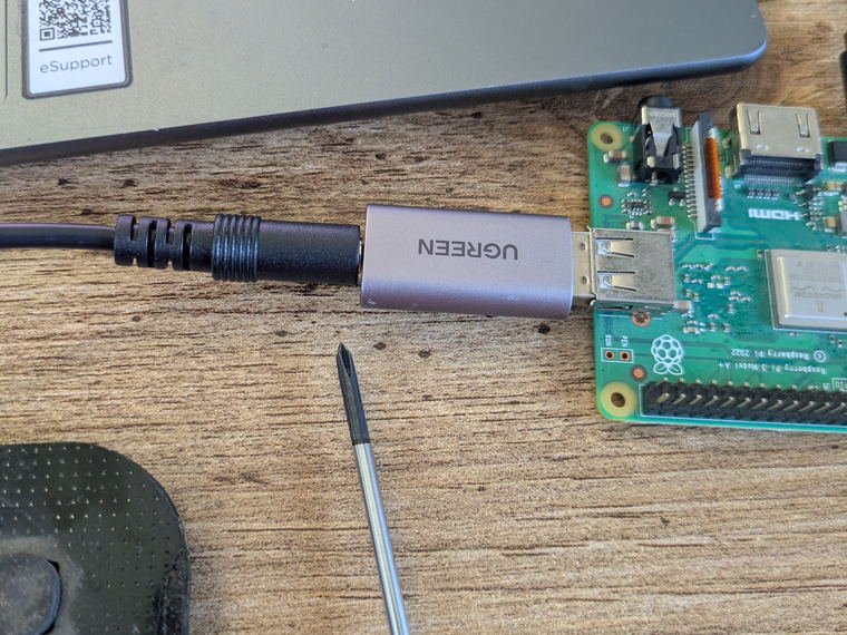 A Raspberry Pi board on a wooden desk next to a small silver USB audio adapter with a headphone cable plugged into it.